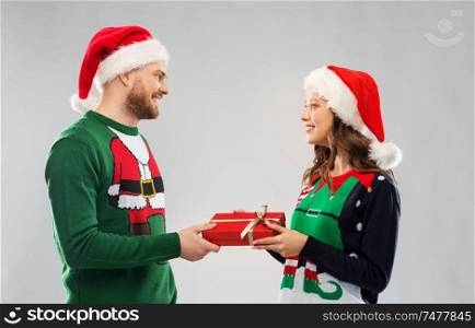 people and holidays concept - happy couple in santa hats with christmas gift at ugly sweater party. happy couple in christmas sweaters with gift box