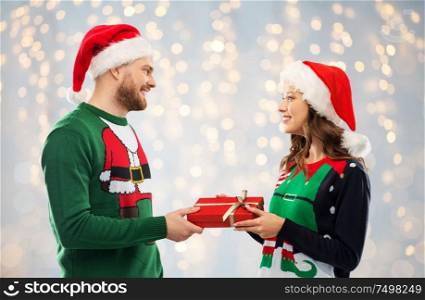 people and holidays concept - happy couple in santa hats with christmas gift at ugly sweater party over festive lights background. happy couple in sweaters with christmas gift