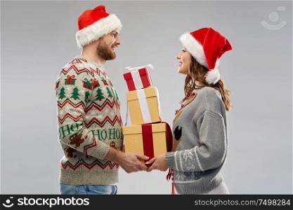 people and holidays concept - happy couple in santa hats with christmas gifts at ugly sweater party. happy couple in christmas sweaters with gifts