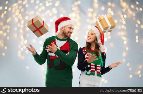 people and holidays concept - happy couple in santa hats with christmas gifts at ugly sweater party over festive lights background. happy couple in ugly sweaters with christmas gifts