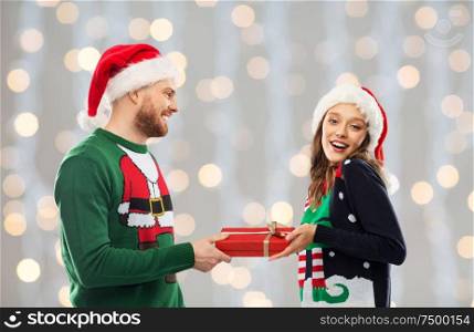 people and holidays concept - happy couple in santa hats with christmas gift at ugly sweater party over festive lights background. happy couple in christmas sweaters with gift box