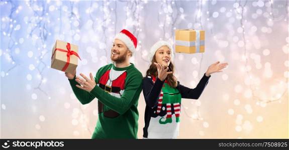 people and holidays concept - happy couple in santa hats with christmas gifts at ugly sweater party over festive lights background. happy couple in christmas sweaters with gifts