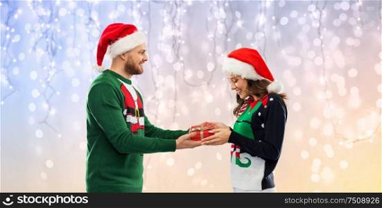 people and holidays concept - happy couple in santa hats with christmas gift at ugly sweater party over festive lights background. happy couple in christmas sweaters with gift box