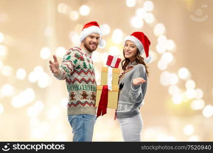 people and holidays concept - happy couple in santa hats with christmas gifts at ugly sweater party over festive lights background. happy couple in christmas sweaters with gifts