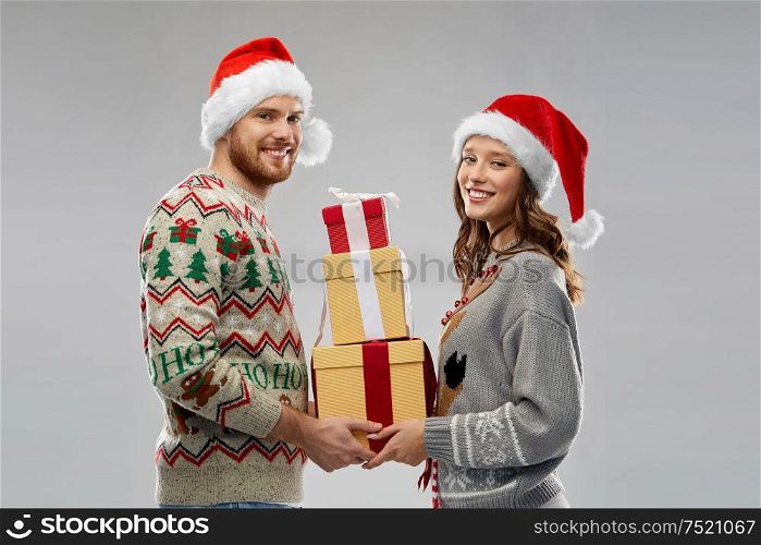 people and holidays concept - happy couple in santa hats with christmas gifts at ugly sweater party. happy couple in christmas sweaters with gifts