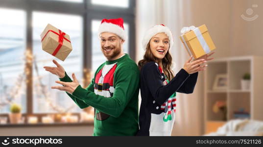 people and holidays concept - happy couple in santa hats with christmas gifts at ugly sweater party over home room background. happy couple in christmas sweaters with gifts