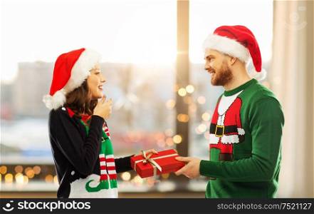 people and holidays concept - happy couple in santa hats with christmas gift at ugly sweater party over home room background. happy couple in christmas sweaters with gift box