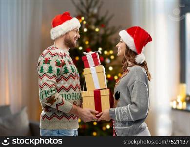 people and holidays concept - happy couple in santa hats with christmas gifts at ugly sweater party over christmas tree lights and home background. couple in christmas sweaters with gifts at home