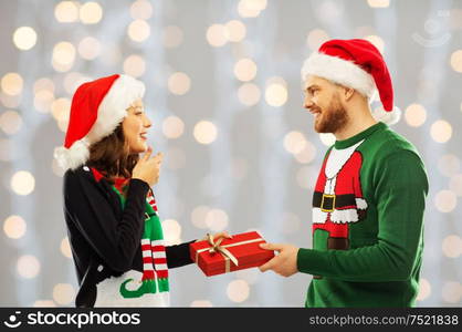 people and holidays concept - happy couple in santa hats with christmas gift at ugly sweater party over festive lights background. happy couple in christmas sweaters with gift box