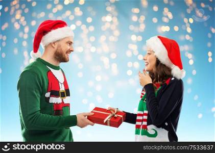 people and holidays concept - happy couple in santa hats with christmas gift at ugly sweater party over festive lights on blue background. happy couple in christmas sweaters with gift box