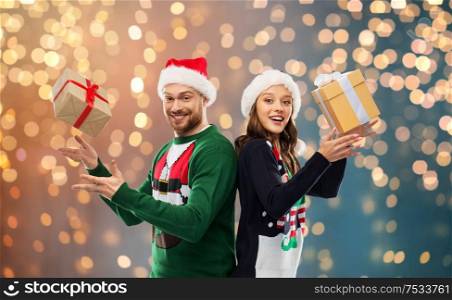 people and holidays concept - happy couple in santa hats with christmas gifts at ugly sweater party over festive lights background. happy couple in christmas sweaters with gifts