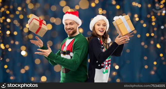 people and holidays concept - happy couple in santa hats with christmas gifts at ugly sweater party over festive lights on dark night background. happy couple in christmas sweaters with gifts