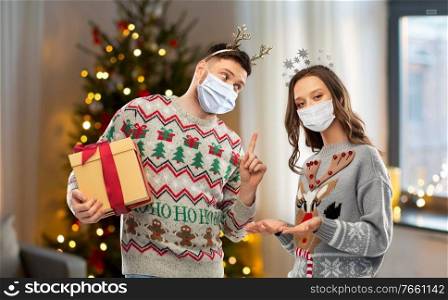 people and holidays concept - happy couple wearing face protective medical mask for protection from virus disease with christmas gift in sweaters at home. couple in masks, christmas sweaters with gifts