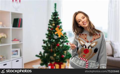 people and holidays concept - happy young woman in jumper with reindeer pattern holding gingerbread accessory at ugly sweater party over christmas tree on home background. woman in christmas sweater with party accessory
