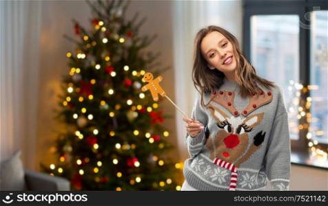 people and holidays concept - happy young woman in jumper with reindeer pattern holding gingerbread accessory at home with christmas tree. woman in christmas sweater with party accessory