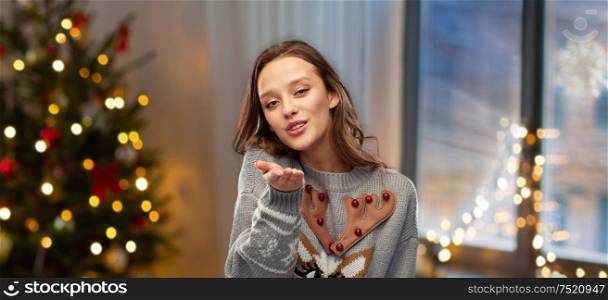 people and holidays concept - happy young woman wearing ugly sweater with reindeer pattern sending air kiss over home and christmas tree lights on background. woman in ugly christmas sweater sending air kiss