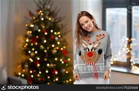 people and holidays concept - happy young woman wearing ugly sweater with reindeer pattern over home and christmas tree lights on background. woman in christmas sweater with reindeer pattern