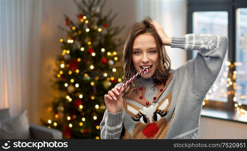 people and holidays concept - happy young woman wearing ugly sweater with reindeer pattern biting candy cane over home and christmas tree lights on background. woman in christmas sweater biting candy cane