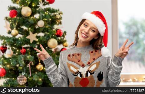 people and holidays concept - happy young woman wearing ugly sweater with reindeer pattern showing peace over christmas tree on background. woman in ugly christmas sweater showing peace sign