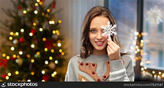 people and holidays concept - happy young woman with snowflake decoration wearing ugly sweater over home and christmas tree lights on background. woman in christmas sweater with snowflake at home