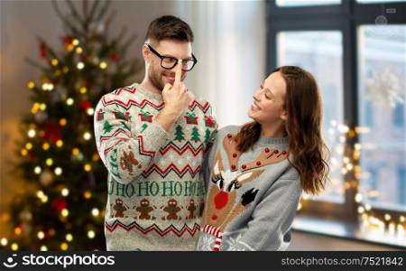 people and holidays concept - portrait of happy couple at ugly sweater party over christmas tree lights and home background. happy couple in christmas ugly sweaters at home