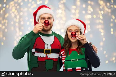 people and holidays concept - portrait of happy couple in santa hats making noses of red christmas balls at ugly sweater party over festive lights background. happy couple in christmas sweaters and santa hats