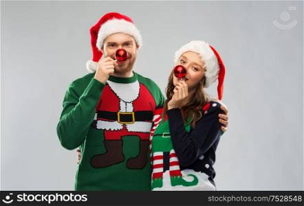 people and holidays concept - portrait of happy couple in santa hats making noses of red christmas balls at ugly sweater party. happy couple in christmas sweaters and santa hats