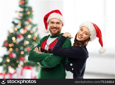 people and holidays concept - portrait of happy couple in santa hats at ugly sweater party over christmas tree lights background. happy couple in christmas sweaters and santa hats
