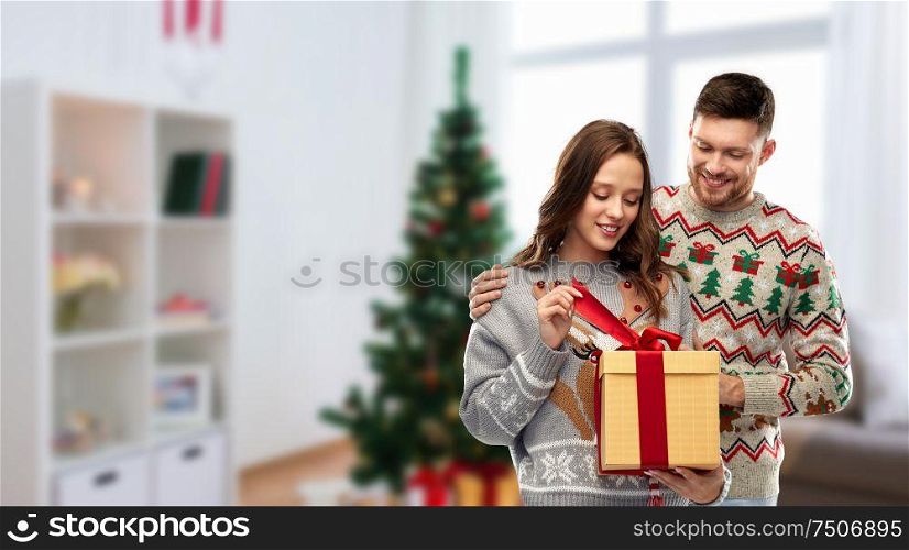 people and holidays concept - portrait of happy couple with christmas gift at ugly sweater party over home room background. happy couple in christmas sweaters with gift box