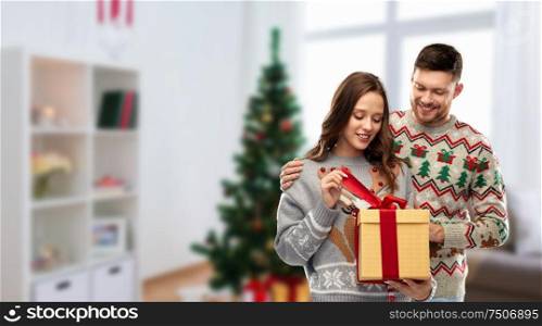 people and holidays concept - portrait of happy couple with christmas gift at ugly sweater party over home room background. happy couple in christmas sweaters with gift box