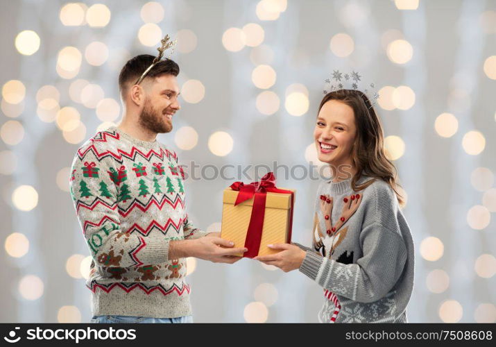 people and holidays concept - portrait of happy couple with christmas gift at ugly sweater party over festive lights background. happy couple in ugly sweaters with christmas gift