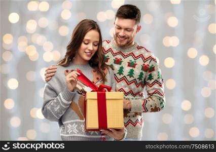 people and holidays concept - portrait of happy couple with christmas gift at ugly sweater party over festive lights background. happy couple in christmas sweaters with gift box