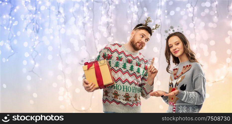people and holidays concept - portrait of happy couple with christmas gift at ugly sweater party over festive lights background. happy couple in ugly sweaters with christmas gift