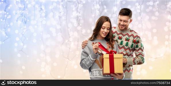 people and holidays concept - portrait of happy couple with christmas gift at ugly sweater party over festive lights background. happy couple in christmas sweaters with gift box