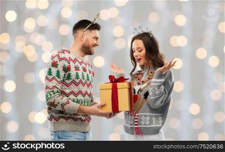 people and holidays concept - portrait of happy couple with christmas gift at ugly sweater party over festive lights background. happy couple in ugly sweaters with christmas gift