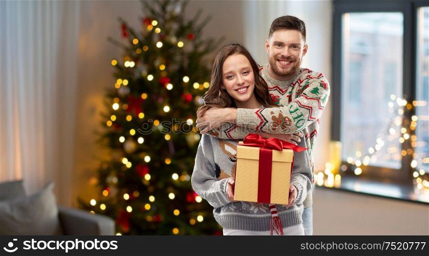 people and holidays concept - portrait of happy couple with christmas gift at ugly sweater party over home background. happy couple in christmas sweaters with gift box