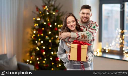 people and holidays concept - portrait of happy couple with christmas gift at ugly sweater party over home background. happy couple in christmas sweaters with gift box