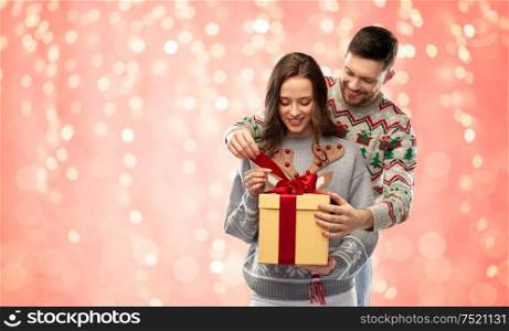 people and holidays concept - portrait of happy couple with christmas gift at ugly sweater party over festive lights on pink coral background. happy couple in christmas sweaters with gift box