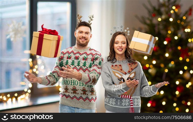 people and holidays concept - portrait of happy couple with christmas gifts at ugly sweater party over home room background. happy couple in christmas sweaters with gift boxes