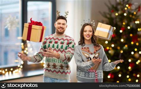 people and holidays concept - portrait of happy couple with christmas gifts at ugly sweater party over home room background. happy couple in christmas sweaters with gift boxes