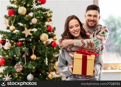 people and holidays concept - portrait of happy couple with gift at ugly sweater party over christmas tree on background. happy couple in christmas sweaters with gift box