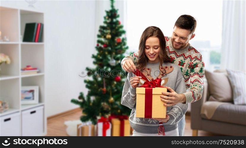 people and holidays concept - portrait of happy couple with gift box at ugly sweater party over christmas tree on home background. happy couple in christmas sweaters with gift box