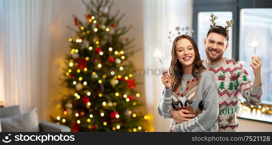 people and holidays concept - portrait of happy couple with sparklers at ugly sweater party over christmas tree lights and home background. happy couple in christmas sweaters with sparklers