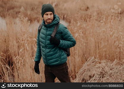People and journey concept. Shot of handsome man dressed in warm jacket and headgear, walks outside, looks attentively aside, poses against field background with copy space on left side for text
