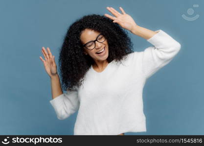 People and joy concept. Optimistic delighted Afro American woman raises hands, dances to loud music, being overjoyed, wears casual white jumper, isolated against blue background, has good mood