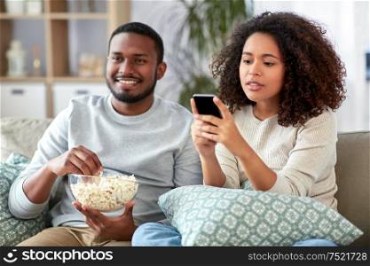 people and leisure concept - african american couple with popcorn and smartphone watching tv at home. african couple with popcorn watching tv at home