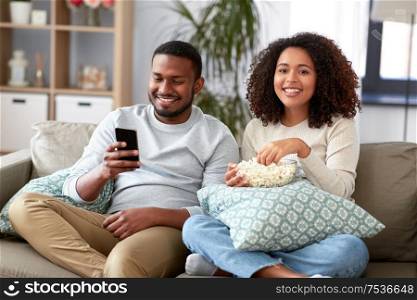 people and leisure concept - african american couple with popcorn and smartphone watching tv at home. african couple with popcorn watching tv at home