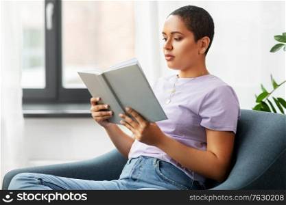 people and leisure concept - african american woman in glasses sitting on sofa and reading book at home. african american woman reading book at home
