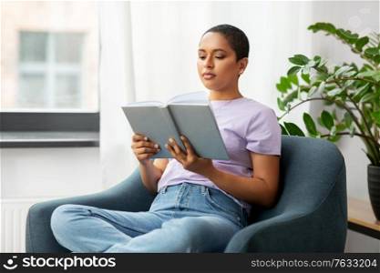 people and leisure concept - african american woman in glasses sitting on sofa and reading book at home. african american woman reading book at home