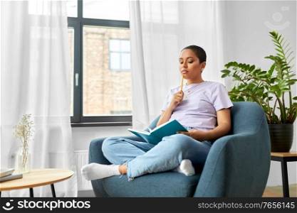 people and leisure concept - african american woman with diary and pencil sitting in chair at home and thinking. african american woman with diary at home